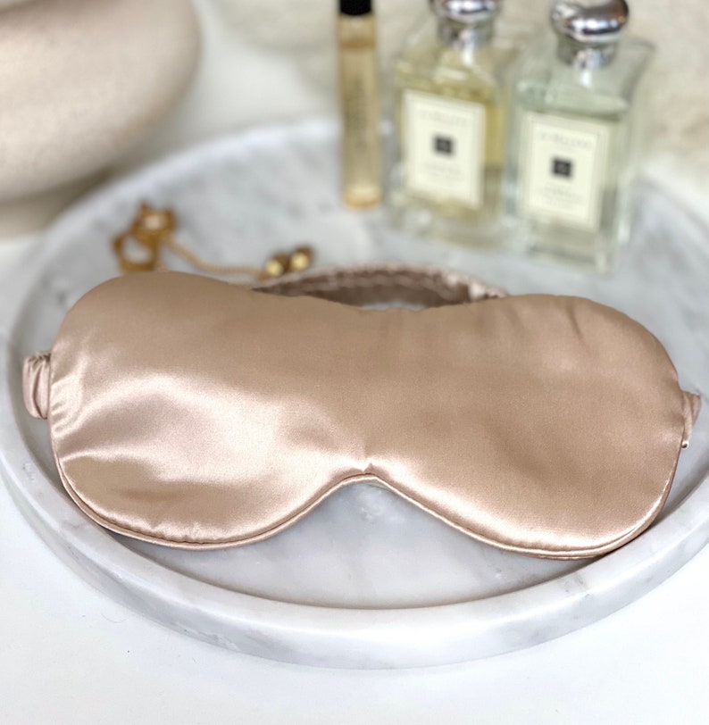 Op de afbeelding: Een lichtbeige satijnen slaapmasker met een dun elastisch bandje.