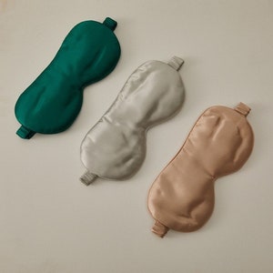 Op de afbeelding: Drie zijden slaapmaskers in verschillende kleuren: groen, lichtgrijs en lichtbruin. De maskers zijn gemaakt van glad, zijdeachtig materiaal en hebben een elastisch bandje om comfortabel om het hoofd te passen.