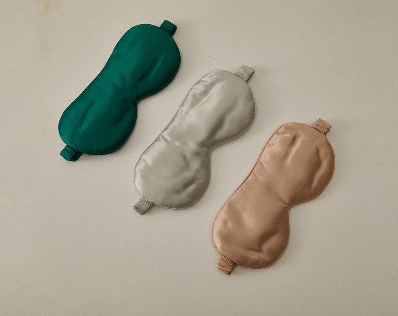Op de afbeelding: Drie zijden slaapmaskers in verschillende kleuren: groen, lichtgrijs en lichtbruin. De maskers zijn gemaakt van glad, zijdeachtig materiaal en hebben een elastisch bandje om comfortabel om het hoofd te passen.