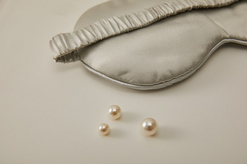 Op de afbeelding: Een grijs zijden slaapmasker met een geplooide elastische band. Drie witte parel kralen zijn verspreid over een lichtgrijs oppervlak.