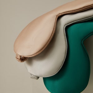 Op de afbeelding: Drie zijden slaapmaskers in verschillende kleuren: lichtbruin, zilver en donkerblauw. De maskers zijn op elkaar gestapeld.