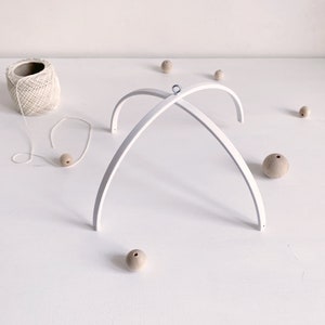 Può includere: Un telaio per mobile in legno bianco con un anello di metallo in cima. Il telaio è composto da due pezzi di legno curvi che si incrociano al centro. Ci sono diverse perline di legno sparse intorno al telaio.