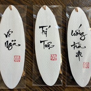 May include: Three white, oval-shaped bookmarks with black calligraphy and red stamps. Each bookmark has a gold tassel and is packaged in clear plastic. The calligraphy appears to be in a foreign language.