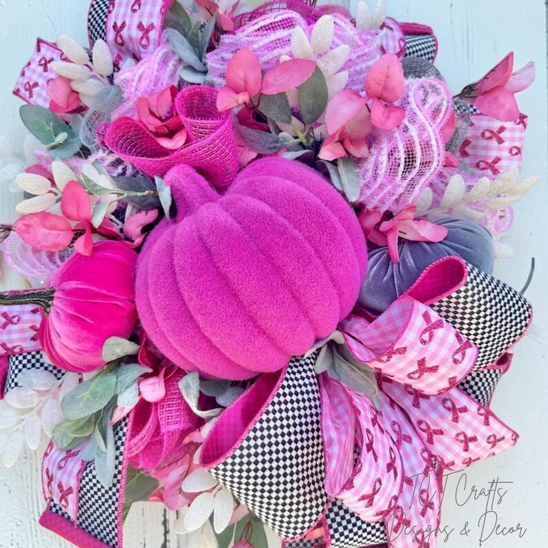 May include: A decorative wreath featuring a large, plush, hot pink pumpkin as the centerpiece. The wreath is adorned with pink and white ribbons, faux flowers, and smaller pumpkins in shades of pink and gray. Black and white checkered ribbon adds contrast.