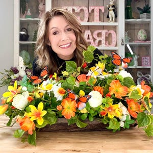 May include: A wooden planter overflowing with vibrant orange, yellow, and white artificial flowers and green leaves. Several orange and black butterfly decorations are interspersed throughout the arrangement. The planter is on a wooden surface.