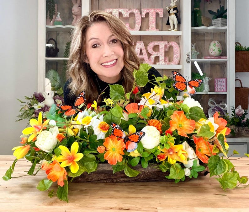 May include: A wooden planter overflowing with vibrant orange, yellow, and white artificial flowers and green leaves. Several orange and black butterfly decorations are interspersed throughout the arrangement. The planter is on a wooden surface.