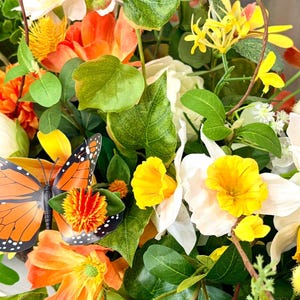 May include: A vibrant floral arrangement featuring orange, yellow, and white flowers, accented by green leaves and a decorative orange and black butterfly. The arrangement includes various flower types and textures.