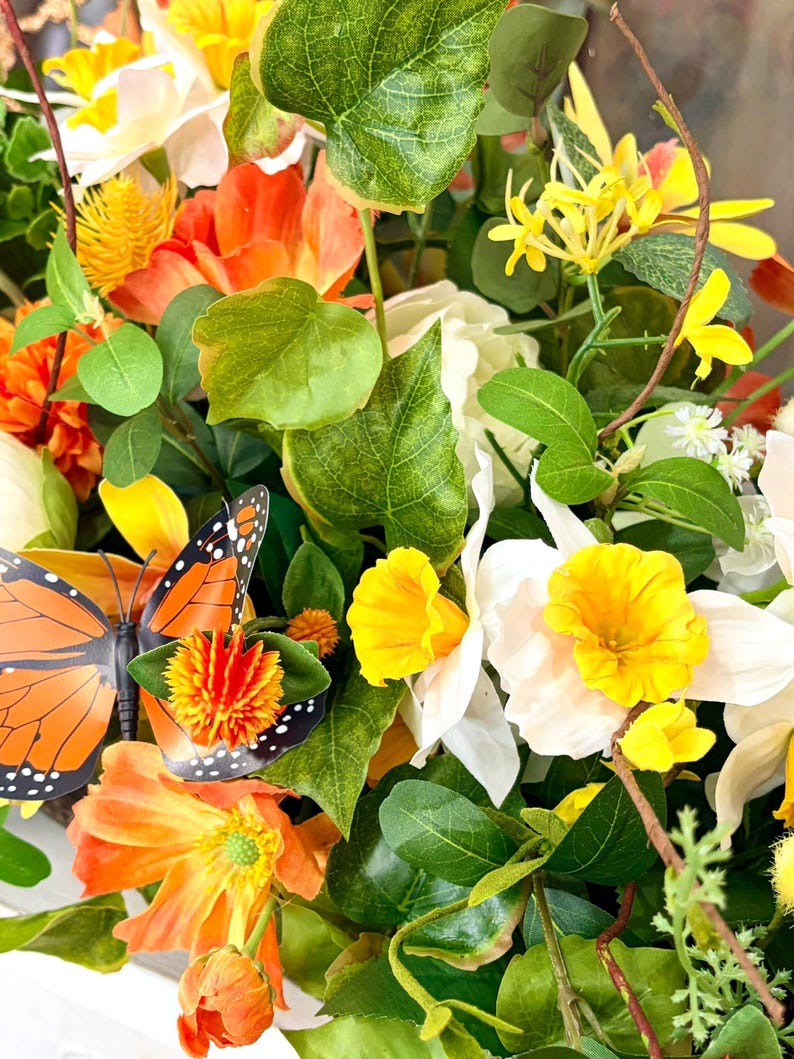 May include: A vibrant floral arrangement featuring orange, yellow, and white flowers, accented by green leaves and a decorative orange and black butterfly. The arrangement includes various flower types and textures.