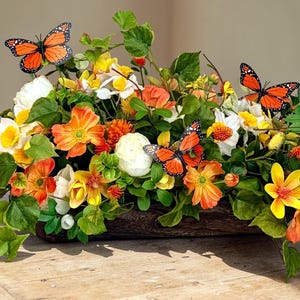May include: A vibrant floral arrangement featuring orange, yellow, and white flowers, accented with green foliage and decorative orange butterflies. The flowers are arranged in a wooden container, creating a natural and colorful display.