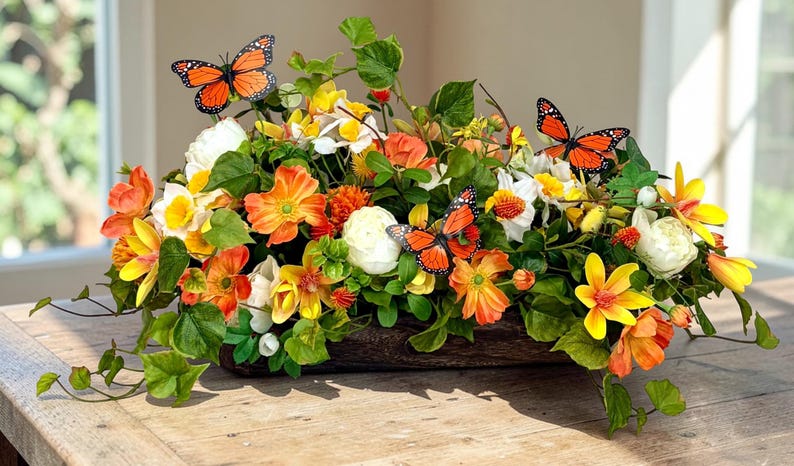May include: A vibrant floral arrangement featuring orange, yellow, and white flowers, accented with green foliage and decorative orange butterflies. The flowers are arranged in a wooden container, creating a natural and colorful display.