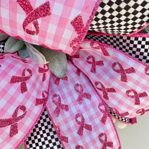 May include: Close-up of a decorative bow featuring pink gingham ribbon printed with pink glitter awareness ribbons. The bow is accented with black and white checkered ribbon and green foliage.
