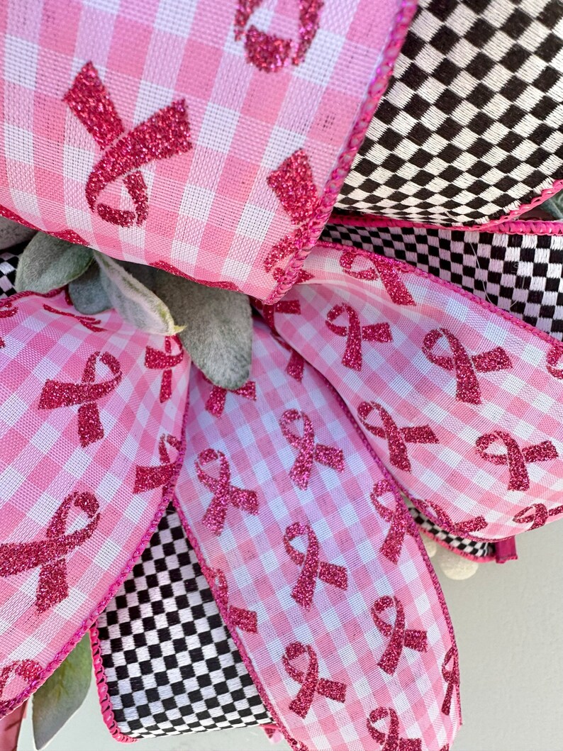 May include: Close-up of a decorative bow featuring pink gingham ribbon printed with pink glitter awareness ribbons. The bow is accented with black and white checkered ribbon and green foliage.