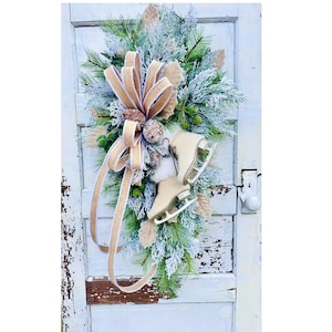 May include: A winter-themed door swag featuring faux evergreen branches, frosted pine needles, and decorative elements. The swag includes a large tan bow, faux berries, and a pair of cream-colored ice skates. The arrangement is designed for seasonal home decor.