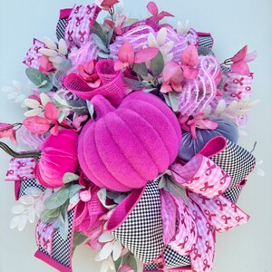 May include: A decorative wreath featuring a large, vibrant pink pumpkin as the centerpiece, surrounded by smaller pumpkins in pink and gray. The wreath incorporates pink ribbons with awareness ribbons, black and white checkered ribbons, and faux foliage.