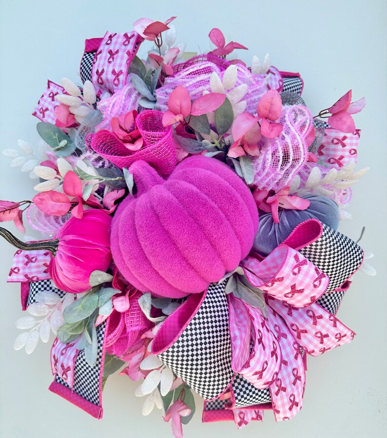 May include: A decorative wreath featuring a large, vibrant pink pumpkin as the centerpiece, surrounded by smaller pumpkins in pink and gray. The wreath incorporates pink ribbons with awareness ribbons, black and white checkered ribbons, and faux foliage.