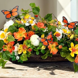 May include: A floral arrangement in a wooden container, featuring orange, yellow, and white flowers with green foliage. Decorative orange and black butterflies are placed throughout the arrangement. The arrangement is on a wooden table.