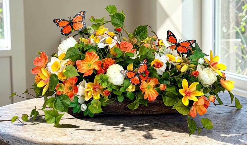 May include: A floral arrangement in a wooden container, featuring orange, yellow, and white flowers with green foliage. Decorative orange and black butterflies are placed throughout the arrangement. The arrangement is on a wooden table.