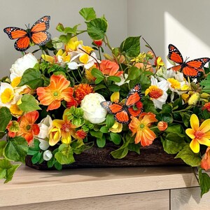 May include: A floral arrangement in a wooden container features orange, yellow, and white flowers with green foliage. Three orange and black butterflies are perched among the flowers. The arrangement sits on a light-colored surface.