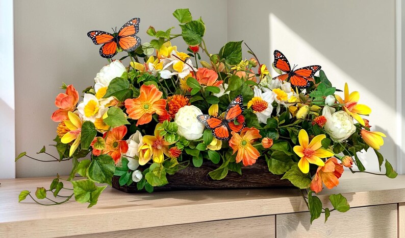 May include: A floral arrangement in a wooden container features orange, yellow, and white flowers with green foliage. Three orange and black butterflies are perched among the flowers. The arrangement sits on a light-colored surface.