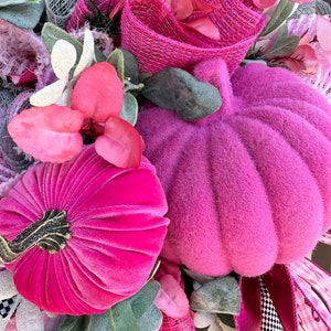 May include: A close-up of a decorative wreath featuring two plush, magenta pumpkins. The wreath is adorned with pink and white ribbons, faux leaves, and various floral elements. The pumpkins have a textured, velvety appearance, and the wreath has a festive, autumnal theme.