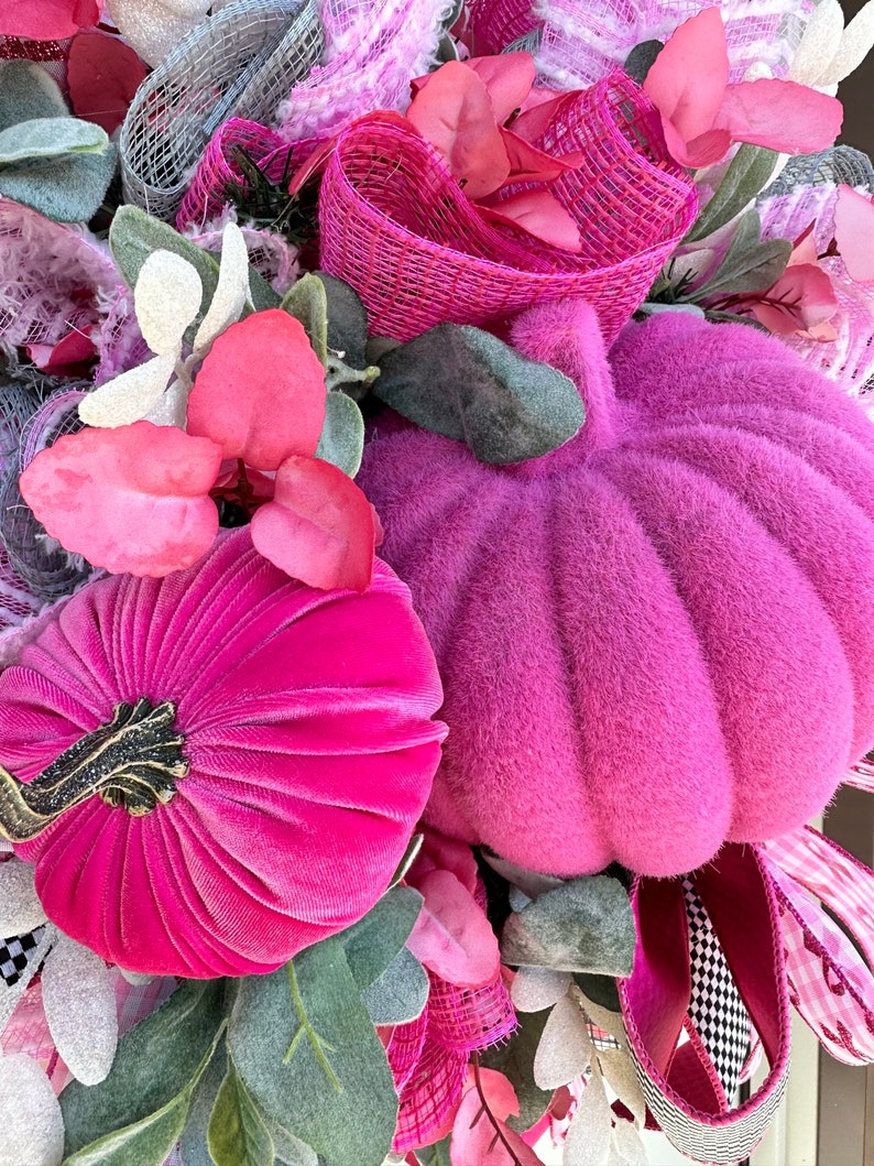 May include: A close-up of a decorative wreath featuring two plush, magenta pumpkins. The wreath is adorned with pink and white ribbons, faux leaves, and various floral elements. The pumpkins have a textured, velvety appearance, and the wreath has a festive, autumnal theme.