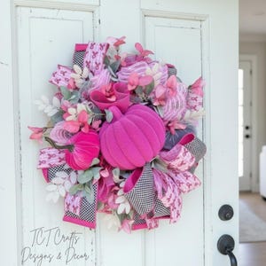 May include: A vibrant pink and white wreath with a pumpkin theme. The wreath features a large, plush pink pumpkin, smaller pink pumpkins, and pink ribbons with a breast cancer awareness ribbon pattern. Accented with pink flowers and greenery.