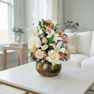 May include: A floral arrangement featuring white lilies, pink roses, and peach-colored carnations in a textured gold-colored metal vase. The vase sits on a white marble-topped coffee table in a living room setting.