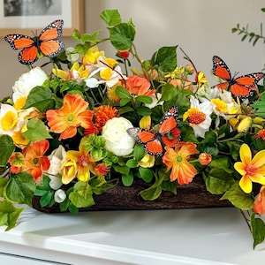 May include: A vibrant floral arrangement in a wooden trough, featuring orange, yellow, and white flowers with green foliage. Decorative orange and black butterflies are interspersed throughout the display. The arrangement sits on a white surface.