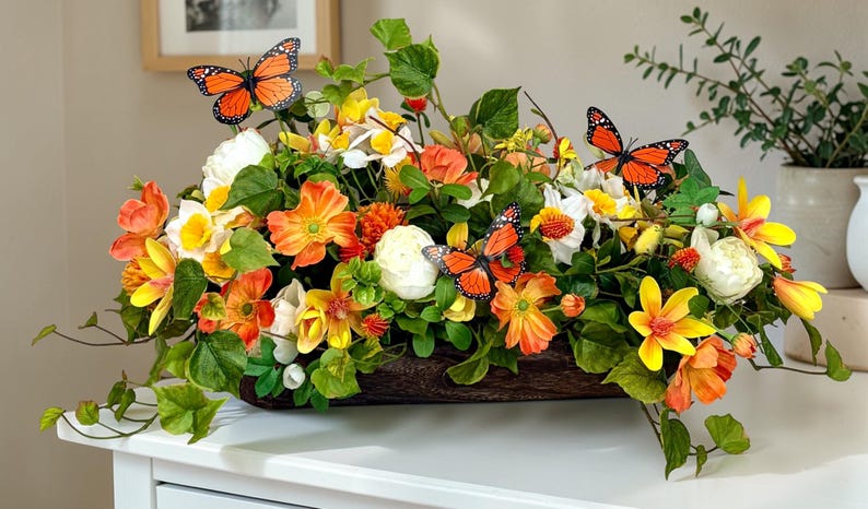 May include: A vibrant floral arrangement in a wooden trough, featuring orange, yellow, and white flowers with green foliage. Decorative orange and black butterflies are interspersed throughout the display. The arrangement sits on a white surface.