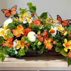 May include: A vibrant floral arrangement in a wooden container, featuring orange, yellow, and white flowers with green foliage. Decorative orange butterflies are interspersed throughout the arrangement, creating a lively and natural aesthetic.