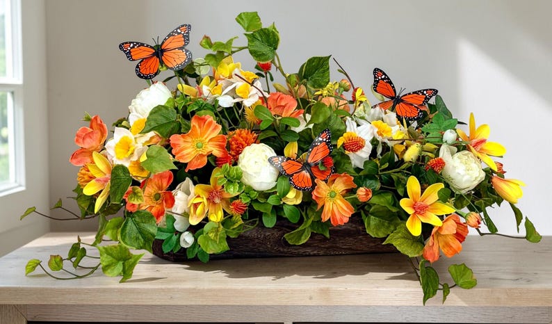 May include: A vibrant floral arrangement in a wooden container, featuring orange, yellow, and white flowers with green foliage. Decorative orange butterflies are interspersed throughout the arrangement, creating a lively and natural aesthetic.