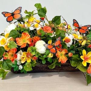 May include: A floral arrangement in a wooden container features orange, yellow, and white flowers with green foliage. Three orange butterflies with black markings are placed among the flowers. The arrangement is on a light-colored surface.