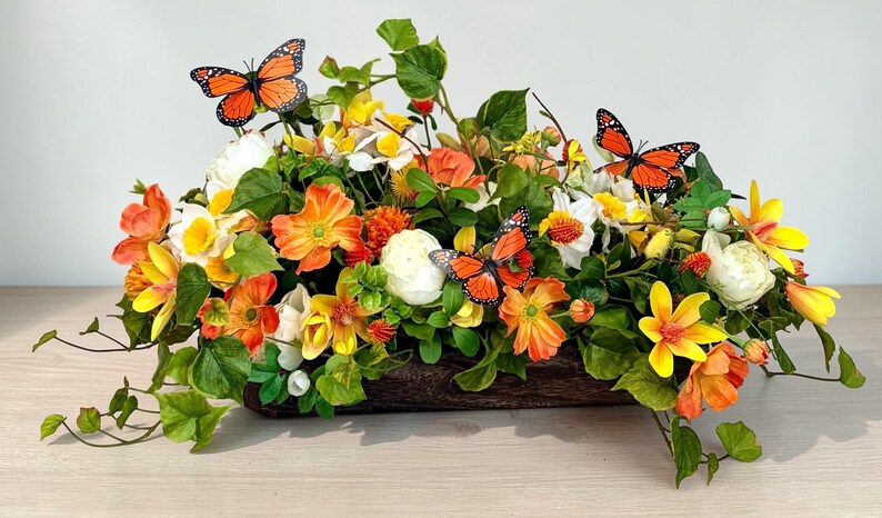 May include: A floral arrangement in a wooden container features orange, yellow, and white flowers with green foliage. Three orange butterflies with black markings are placed among the flowers. The arrangement is on a light-colored surface.
