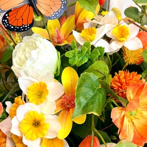 May include: A vibrant floral arrangement featuring a mix of yellow, orange, and white flowers, with green foliage. A decorative butterfly with orange and black wings is positioned above the flowers. The arrangement is a mix of different flower types.
