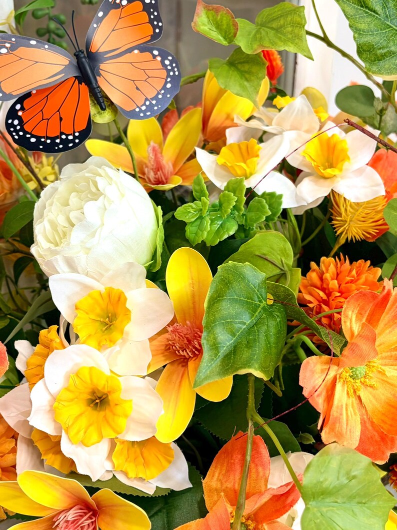 May include: A vibrant floral arrangement featuring a mix of yellow, orange, and white flowers, with green foliage. A decorative butterfly with orange and black wings is positioned above the flowers. The arrangement is a mix of different flower types.