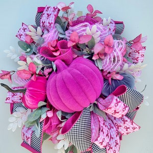 May include: A decorative fall wreath featuring a large, plush, magenta pumpkin as the centerpiece. The wreath is adorned with pink and white ribbons, faux foliage, and smaller velvet pumpkins in shades of pink and gray. The ribbons have a black and white checkered pattern.