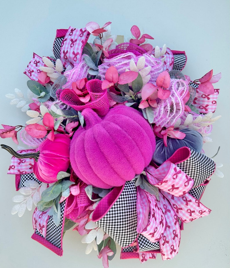 May include: A decorative fall wreath featuring a large, plush, magenta pumpkin as the centerpiece. The wreath is adorned with pink and white ribbons, faux foliage, and smaller velvet pumpkins in shades of pink and gray. The ribbons have a black and white checkered pattern.