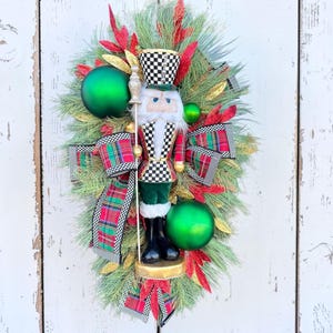 May include: A festive Christmas wreath featuring a nutcracker soldier with a checkered hat and vest, surrounded by green ornaments, red and gold accents, and plaid ribbon. The wreath is set against a white wooden background.