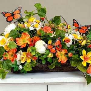 May include: A vibrant floral arrangement in a wooden container, featuring orange, yellow, and white flowers with green foliage. Decorative orange butterflies are interspersed throughout the display, creating a lively and colorful centerpiece.