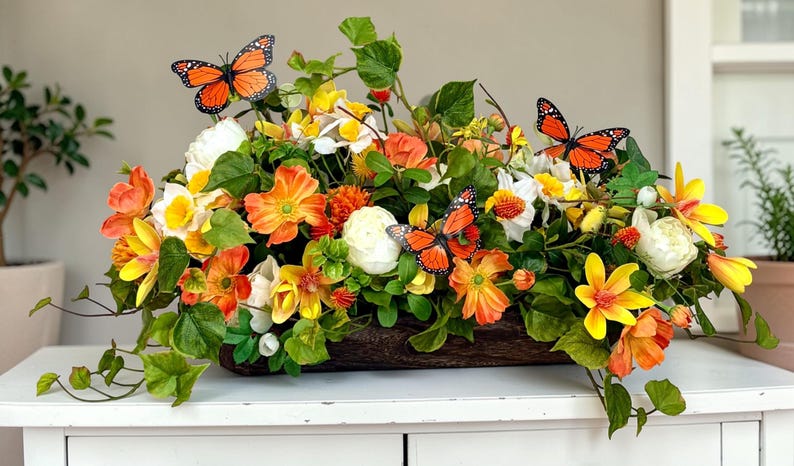 May include: A vibrant floral arrangement in a wooden container, featuring orange, yellow, and white flowers with green foliage. Decorative orange butterflies are interspersed throughout the display, creating a lively and colorful centerpiece.