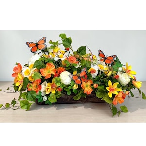 May include: A rectangular floral arrangement featuring a variety of colorful flowers in shades of orange, yellow, and white, with green foliage. Three orange butterflies with black markings are placed throughout the arrangement. The flowers are in a brown wooden container.