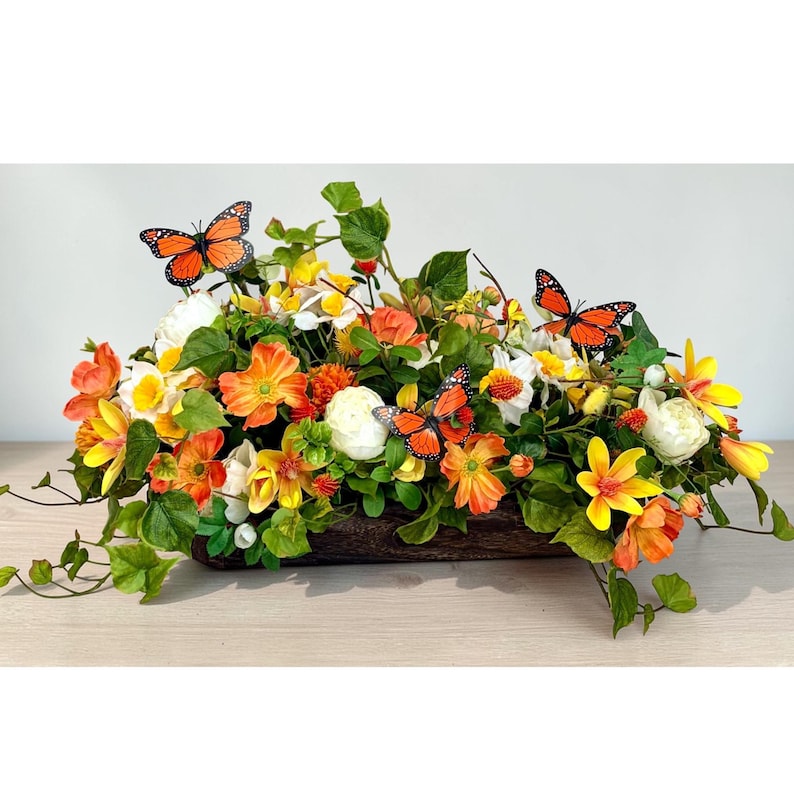 May include: A rectangular floral arrangement featuring a variety of colorful flowers in shades of orange, yellow, and white, with green foliage. Three orange butterflies with black markings are placed throughout the arrangement. The flowers are in a brown wooden container.