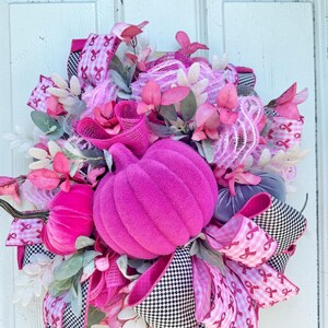 May include: A vibrant, handmade wreath featuring a large, plush, magenta pumpkin as the centerpiece. The wreath is adorned with pink ribbons, faux flowers, and greenery, with accents of black and white checkered ribbon. A festive autumn decoration.