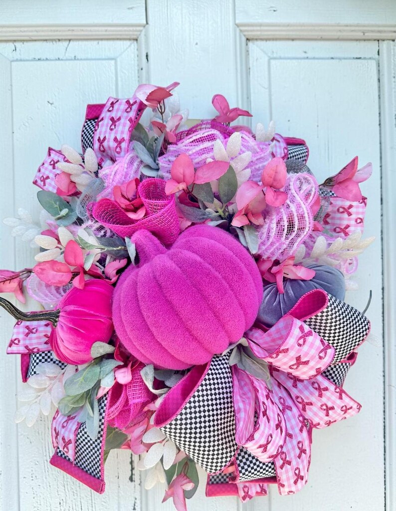 May include: A vibrant, handmade wreath featuring a large, plush, magenta pumpkin as the centerpiece. The wreath is adorned with pink ribbons, faux flowers, and greenery, with accents of black and white checkered ribbon. A festive autumn decoration.