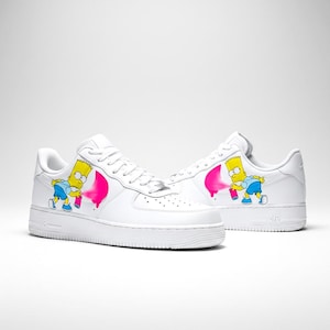 Puede incluir: Zapatillas blancas con un diseño de Bart Simpson rociando pintura rosa. Los zapatos tienen un diseño clásico con cordones y una puntera perforada. El personaje de dibujos animados es amarillo, azul y rosa.