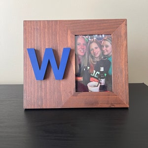 May include: A brown wooden picture frame with a blue letter "W" attached to the front. The frame holds a photo of three women smiling at the camera.