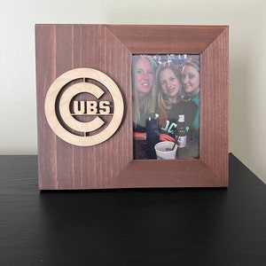 May include: A brown wooden picture frame with a cutout of the Chicago Cubs logo. The frame has a photo of three women in it.