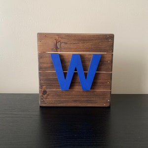 May include: A wooden sign with the letter "W" painted in a bright blue color. The sign is made of reclaimed wood and has a rustic look.