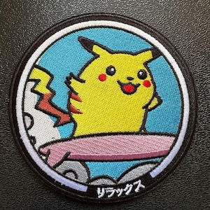 Peut inclure: Un écusson brodé circulaire représentant un personnage Pikachu jaune aux joues rouges, debout sur une planche de surf rose sur un fond bleu clair. Le motif est entouré d'une bordure noire avec des caractères japonais blancs en bas.