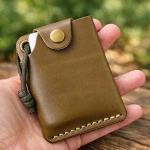 May include: An olive green leather card holder featuring a brass snap button closure, visible light-colored stitching along the bottom edge, and a matching olive green paracord lanyard attached through a reinforced metal eyelet on the side.